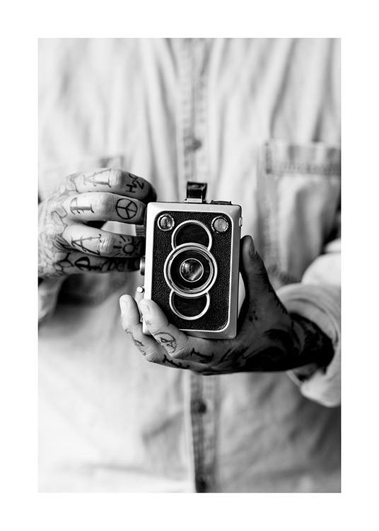  – Photographie en noir et blanc d’un vieil appareil photo tenu par un homme aux mains tatouées