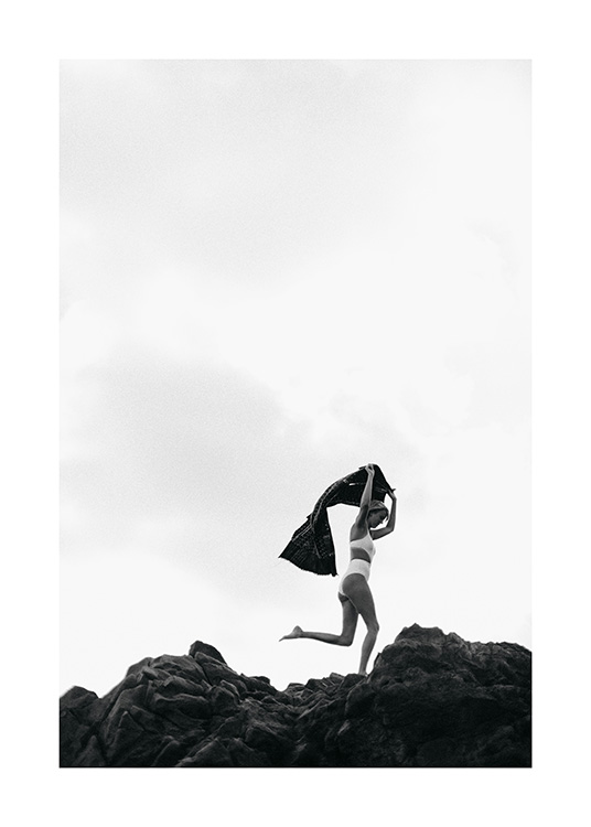  - Photographie en noir et blanc d'une femme en bikini tenant une serviette au-dessus de sa tête et courant sur des rochers