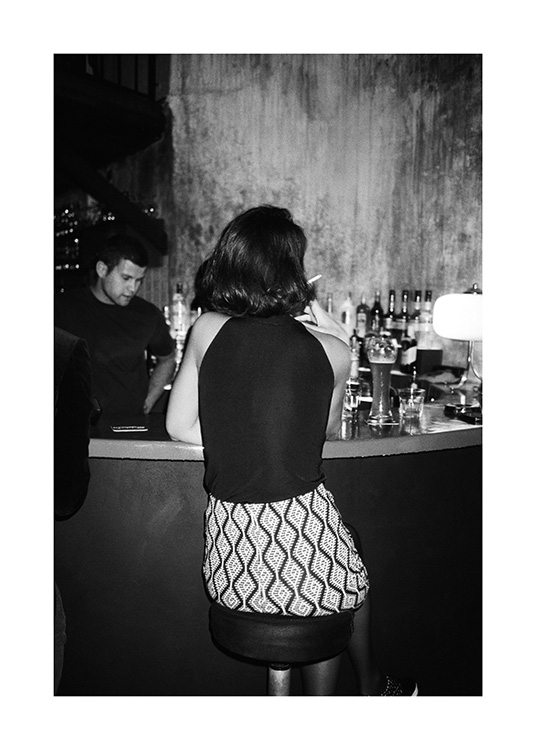  – Photographie en noir et blanc d’une femme assise au comptoir d’un bar, fumant une cigarette