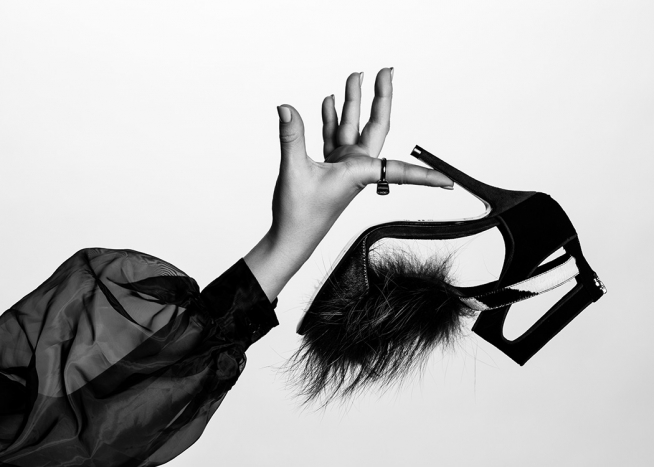  – Photographie en noir et blanc d’une chaussure à talon haut avec de la fourrure sur le bout, tenue par une femme dans un chemisier bouffant très fin