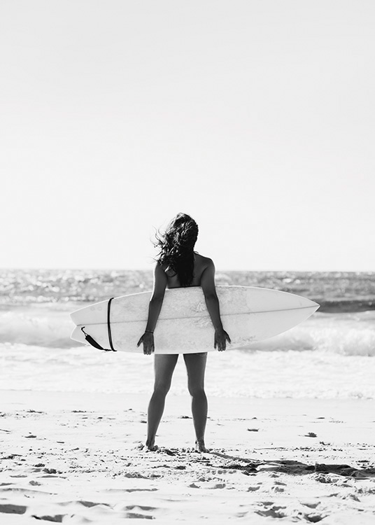  – Photographie en noir et blanc d’une fille tenant une planche de surf derrière elle avec l’océan à l’arrière-plan