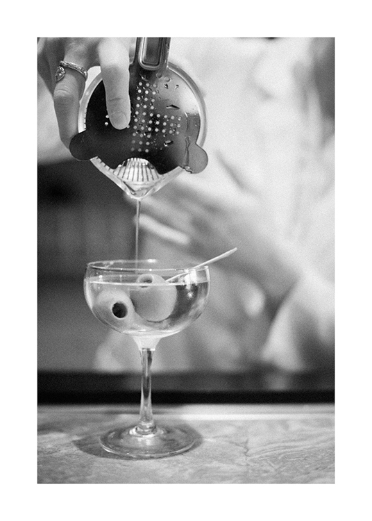  – Photographie en noir et blanc d’un cocktail au martini versé à partir d’un shaker, avec des olives dans le verre