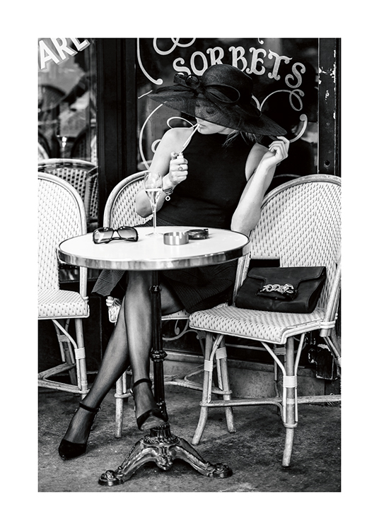  – Photographie en noir et blanc d’une femme assise à la terrasse d’un café, portant un chapeau et allumant une cigarette