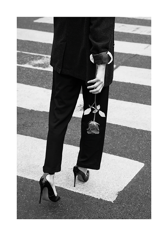 – Photographie en noir et blanc d’une femme marchant sur un passage piéton dans un tailleur pantalon et des talons, tenant une rose