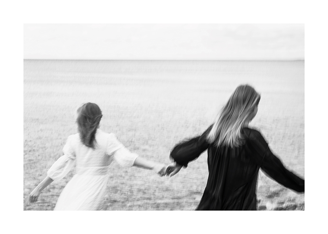  – Photographie en noir et blanc d’un couple de femmes courant à travers champ tout en se donnant la main