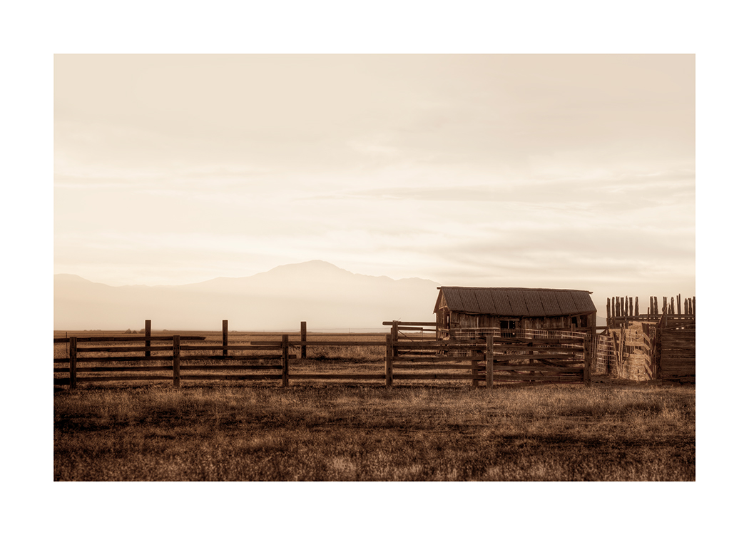 – Photographie d'une grange et de clôtures en bois dans un champ marron, avec des montagnes et un ciel clair à l’arrière-plan